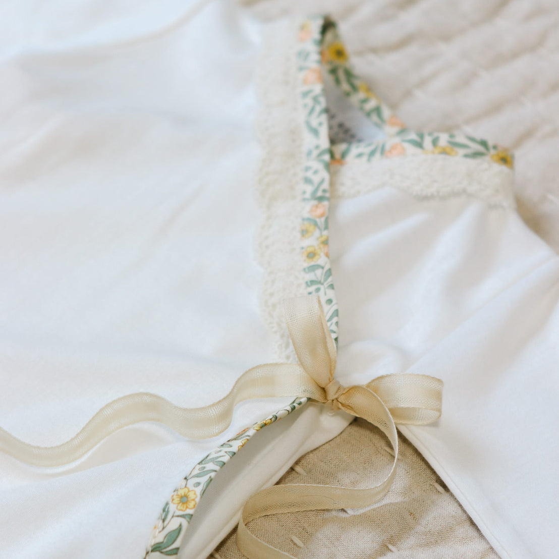Close up flat lay photo of the Lillian Layette. Details show the cotton ribbon that secures the kimo style top and trim in a yellow floral cotton with scalloped champagne lace. 