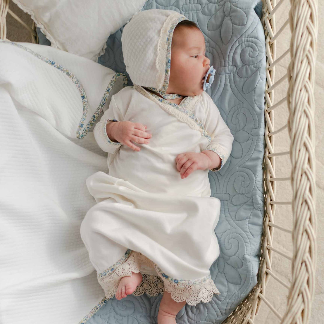 Newborn in a soft cotton layette gown with floral trim and scalloped lace, paired with a quilted bonnet, resting in a wicker bassinet.