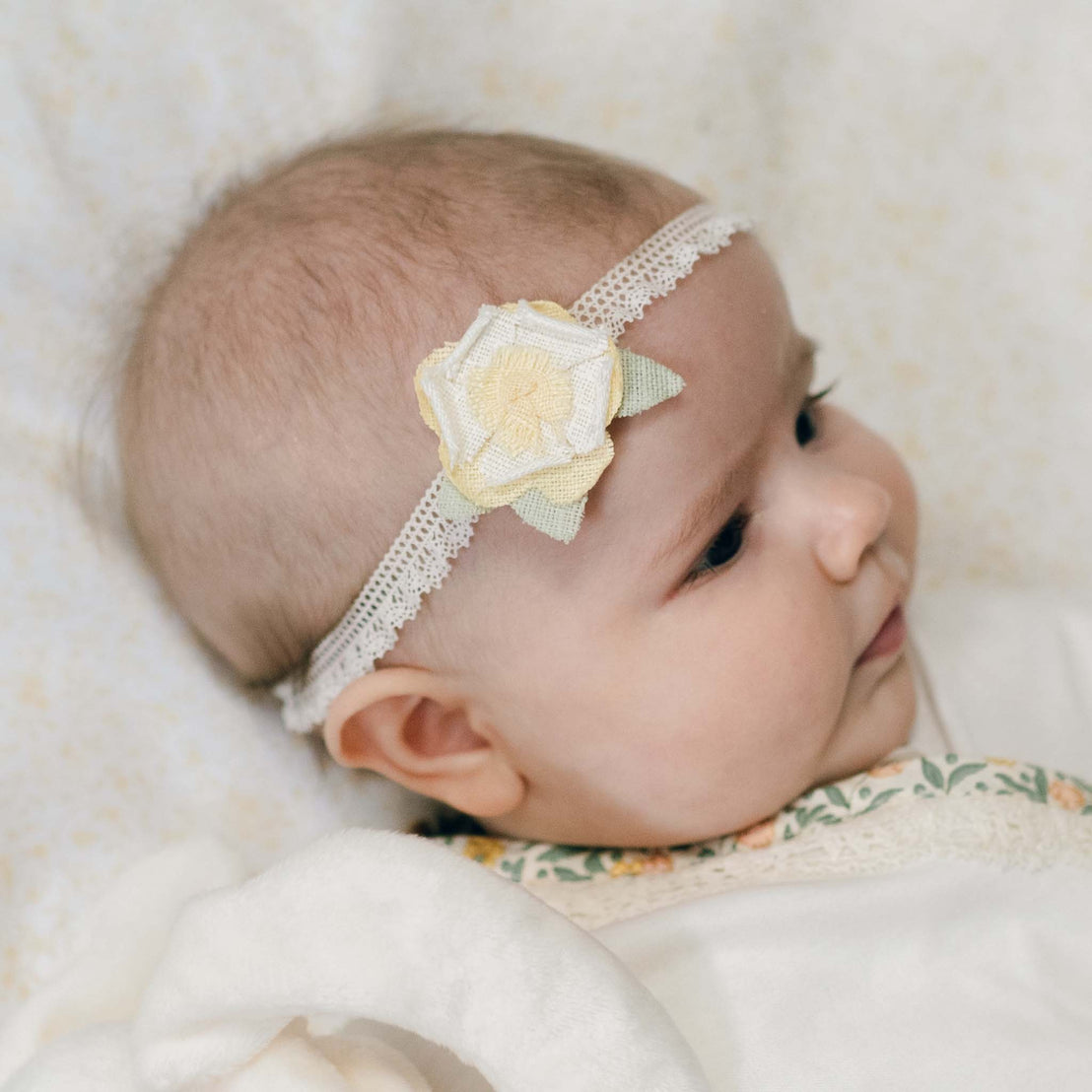Newborn baby wearing the Lillian Linen Flower Headband against a light-colored background. Details of linen flower headband show a yellow flower with stretch lace. Perfect for unique baby gifts.