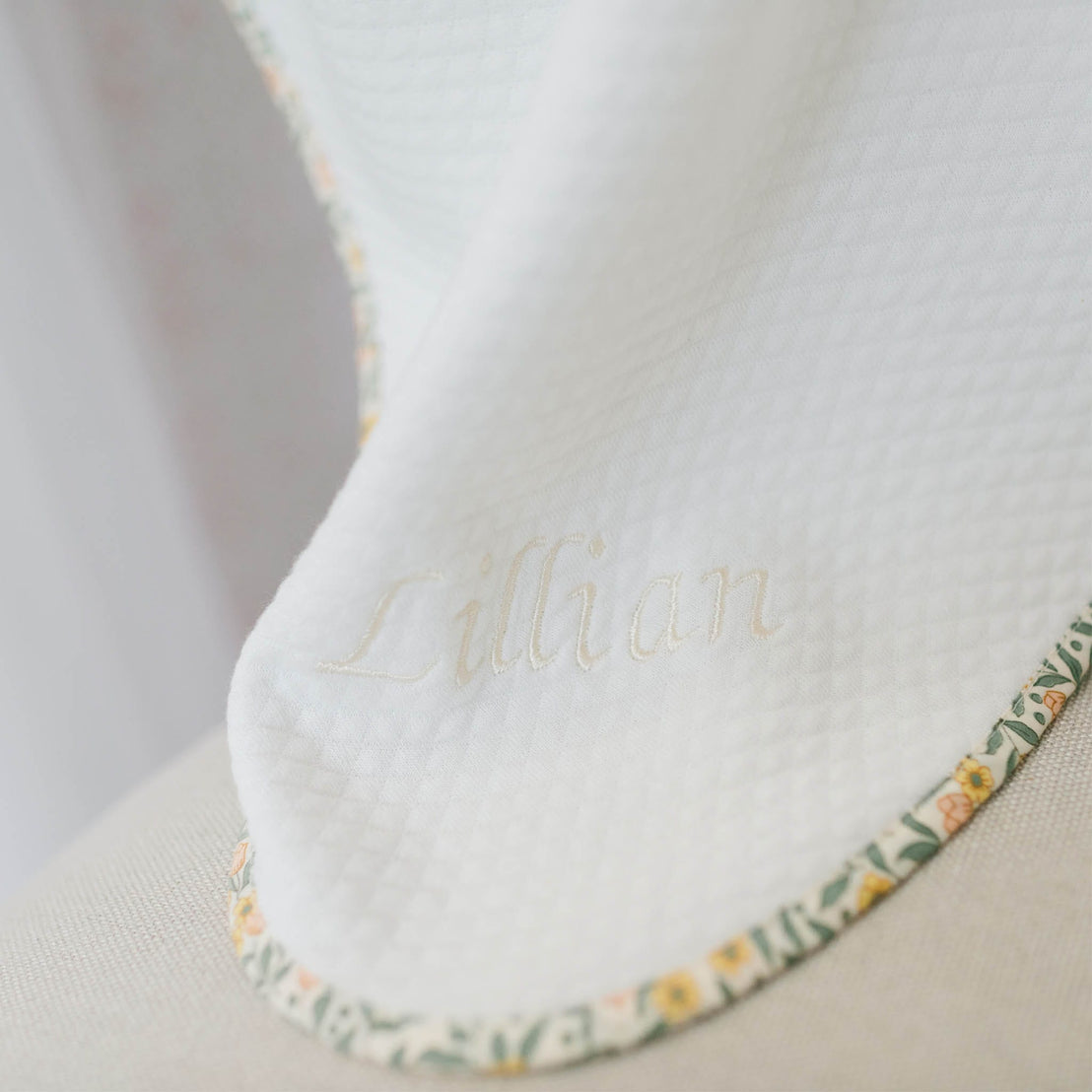 Close up flat lay photo of the Lillian Personalized Blanket on a beige background. Details show yellow floral trim and the name "Lillian" embroidered on the corner of the blanket. Perfect for baptism gift from godparents.