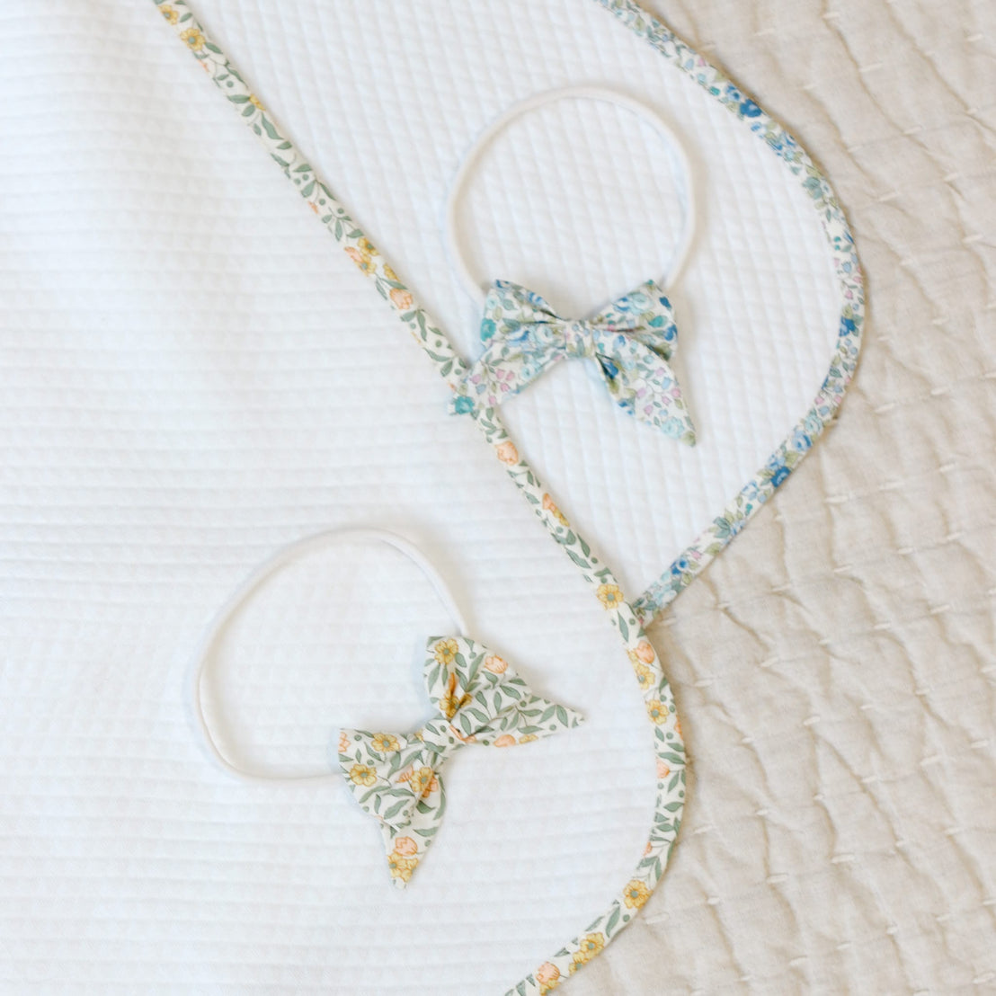 Flat lay photo comparison of the the blue and yellow Lillian Personalized Blanket with matching Headband Bows on a beige background. Details show both the yellow and blue floral fabric on the blanket and bows.