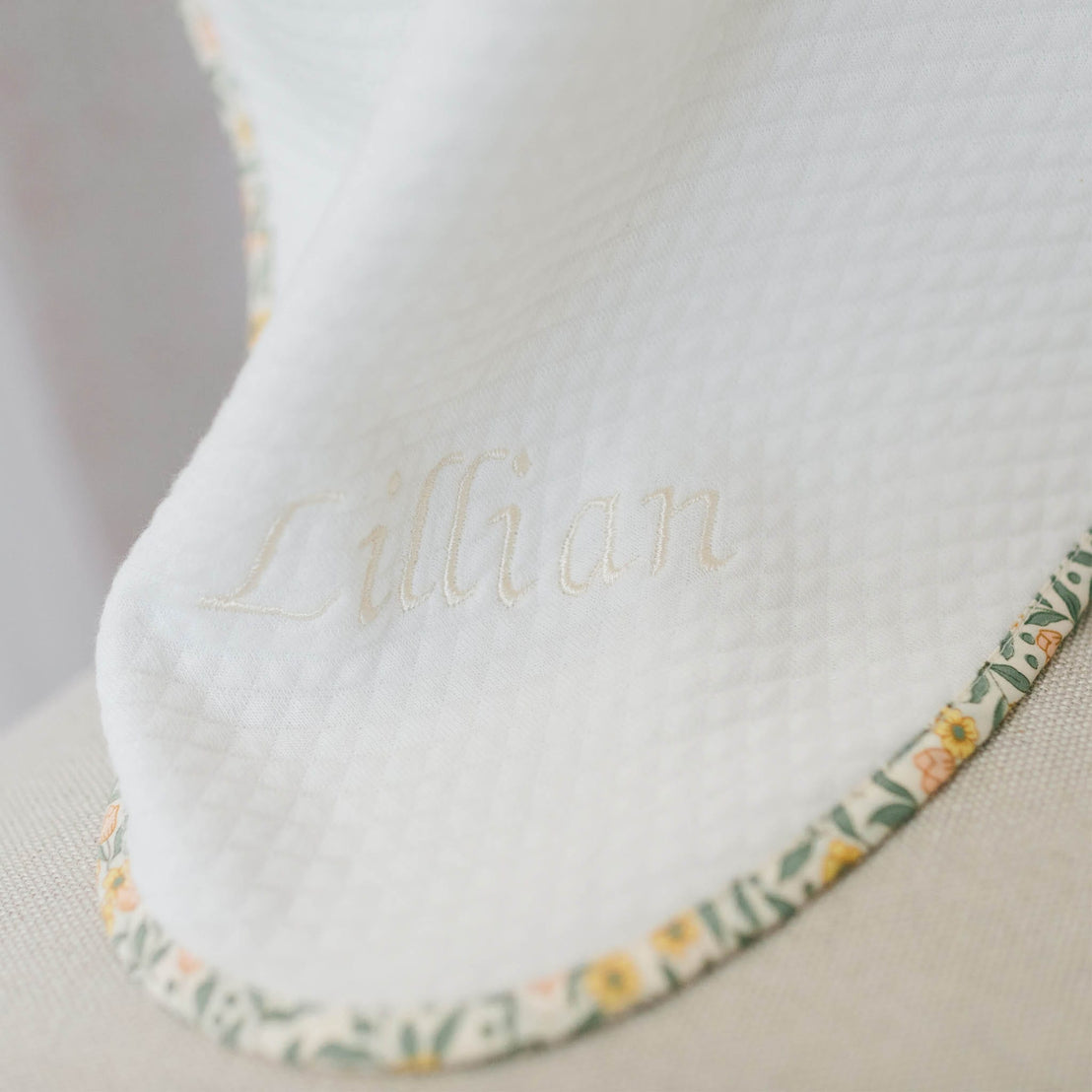 Close up flat lay photo of the Lillian Personalized Blanket on a beige background. Details show yellow floral trim and the name "Lillian" embroidered on the corner of the blanket.