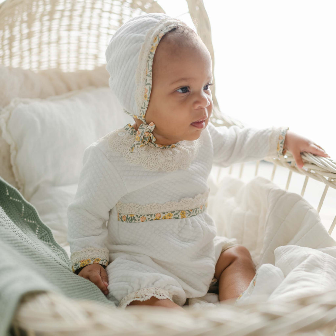 Baby in the Petite Fleur Yellow Romper and bonnet, sitting in a white bassinet with soft blankets.