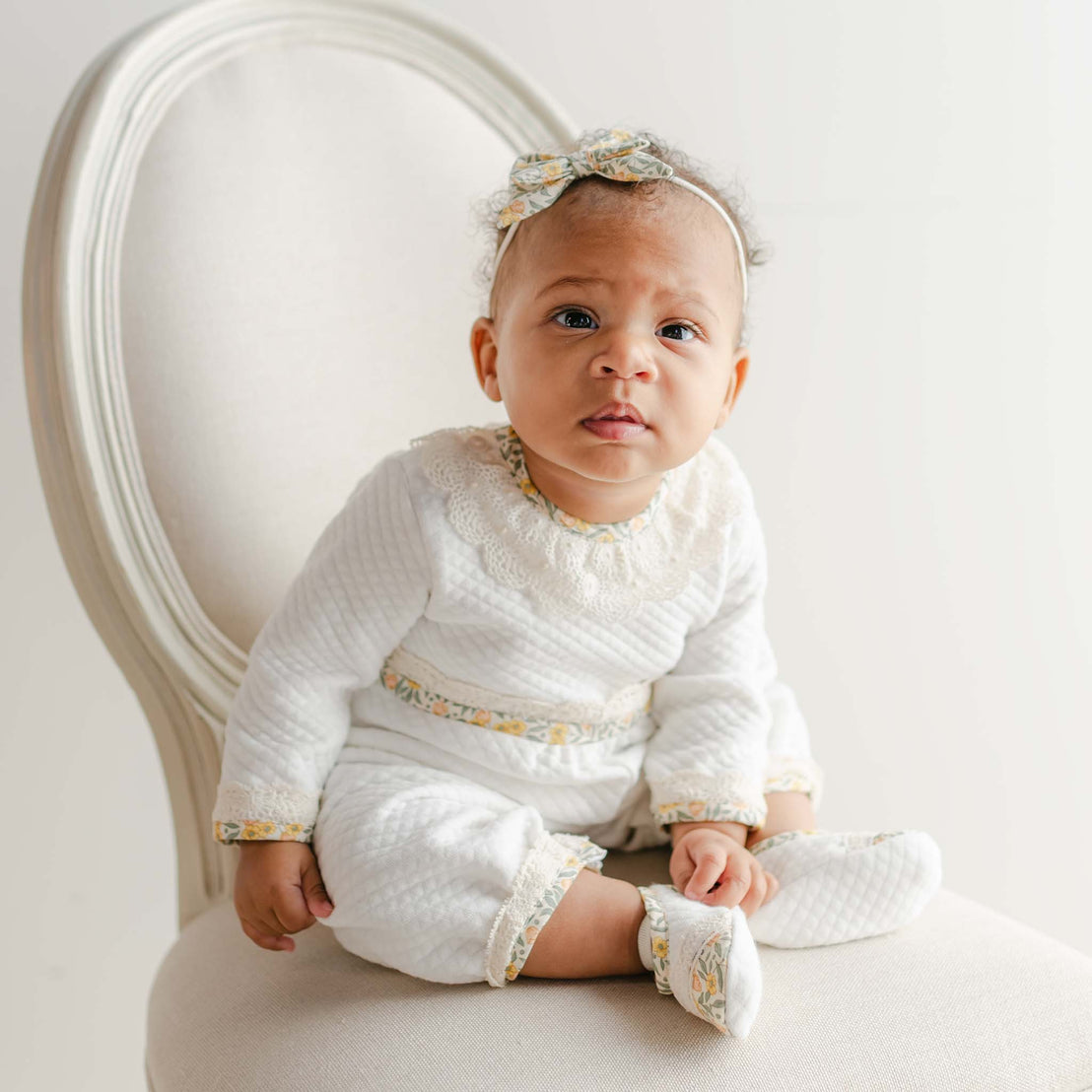 Baby wearing the Petite Fleur Yellow Romper with lace collar, floral trim, and matching headband on a cream chair.