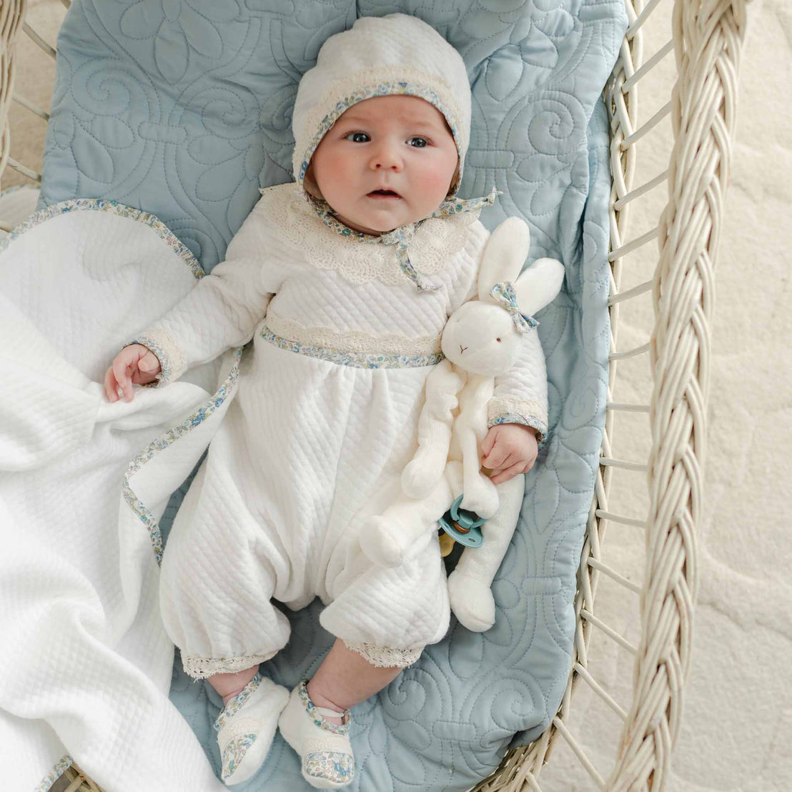 Baby in the Petite Fleur Blue Romper with matching bonnet and bunny plush, resting in a bassinet.