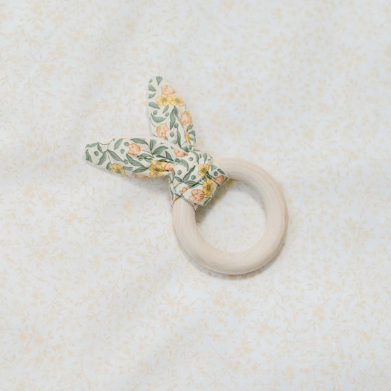 Flat lay photo of the Lillian Wooden Teether Ring against a floral patterned background. Details show maple wood ring with a yellow floral cotton tie. Perfect for unique baby gifts.