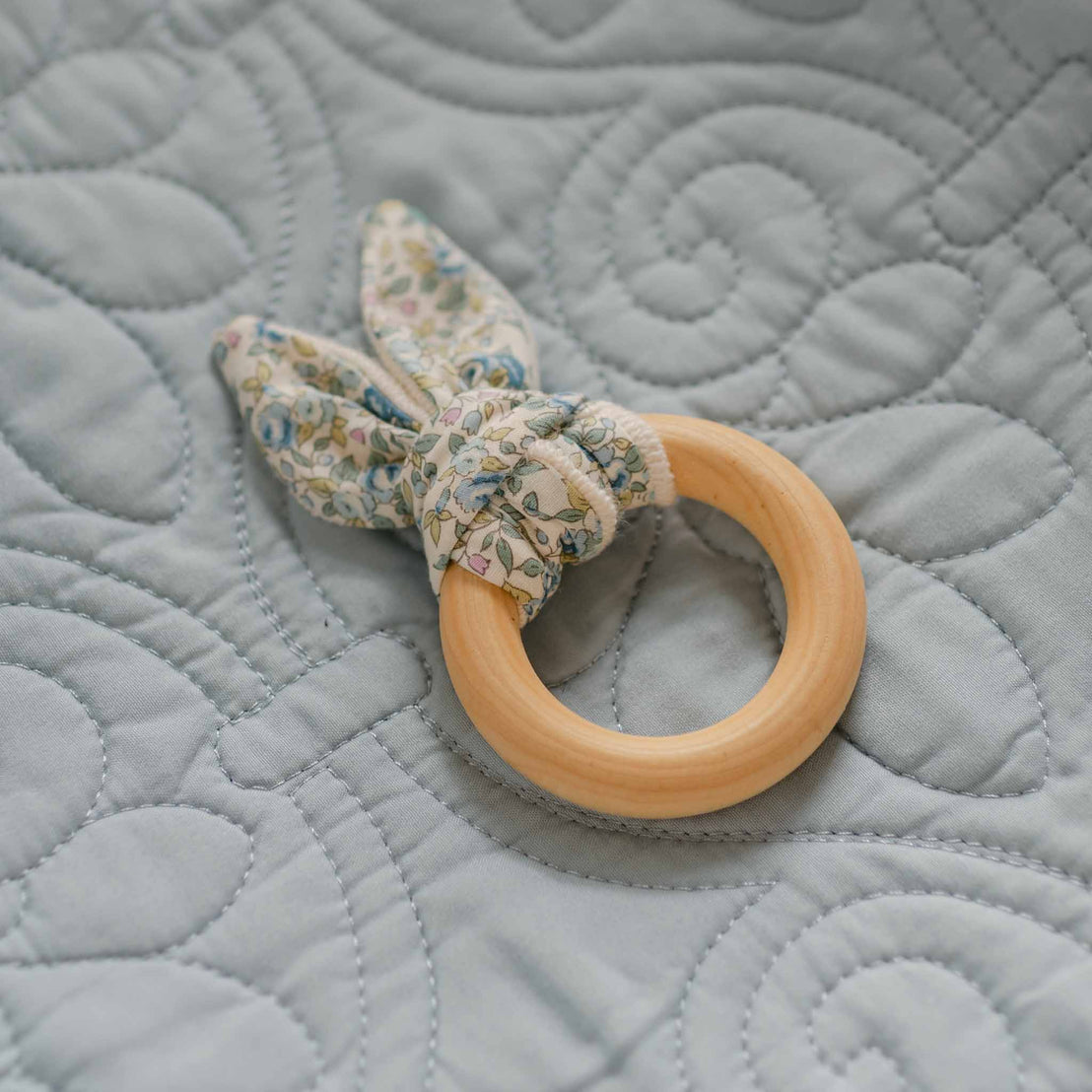 The Petite Fleur Blue Wooden Teether Ring with a floral cotton fabric tie, displayed on a soft quilted blue blanket.