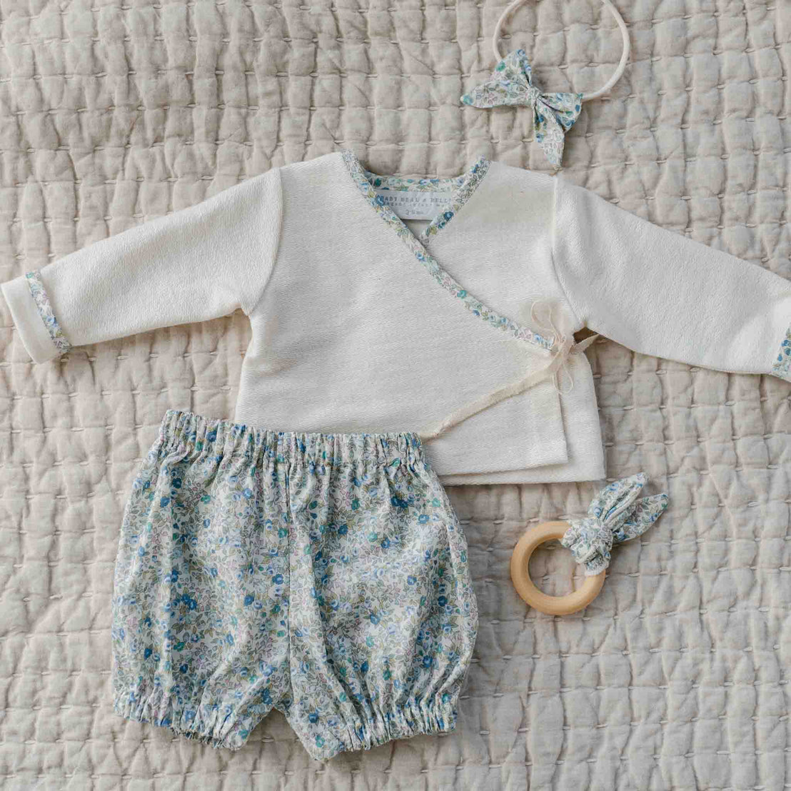 The Petite Fleur Blue Wrap Top & Bloomers set with floral trim, shown with a matching bow headband and wooden teether.