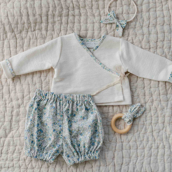 The Petite Fleur Blue Wrap Top & Bloomers set with floral trim, shown with a matching bow headband and wooden teether.