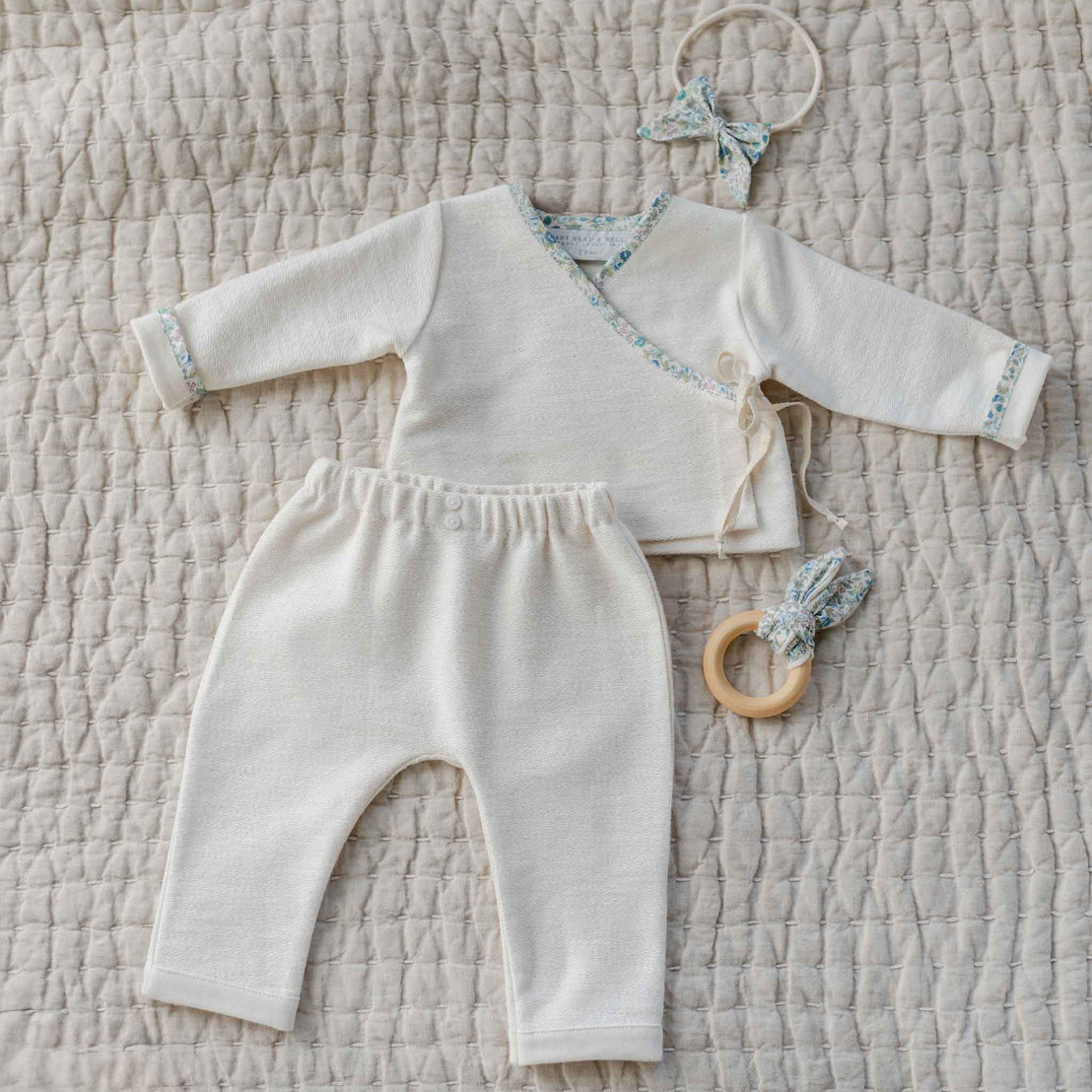 The Petite Fleur Blue Wrap Top & Pants with floral trim, shown with matching bow headband and wooden teether.

