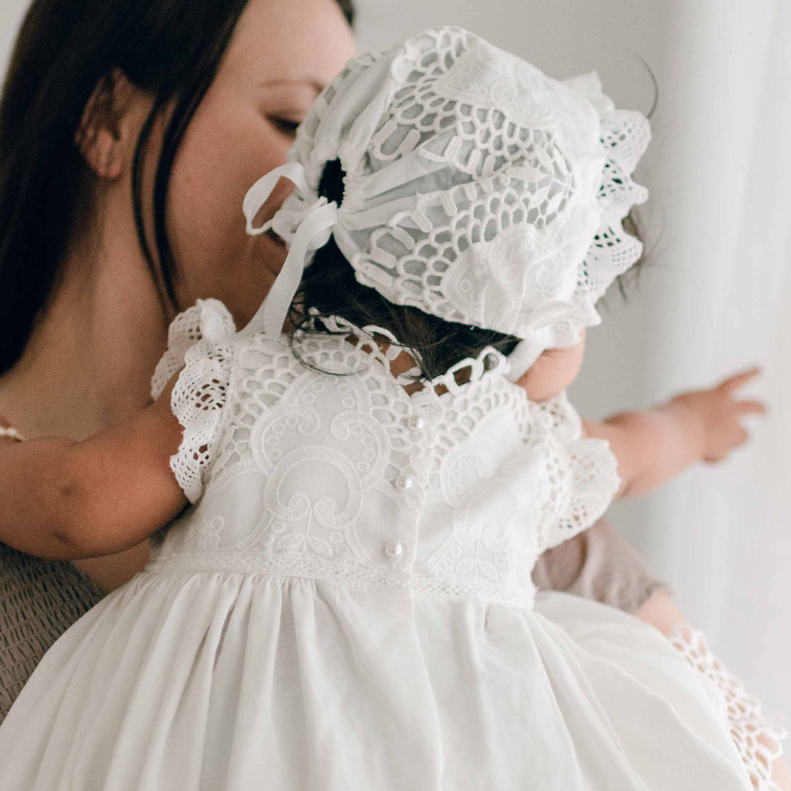 Back view of a baby wearing the Lily Bonnet and matching dress. The baby points to something off to the side as she sits in her mom's arms who appears in the background.