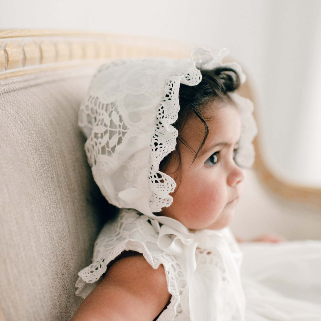 Side profile of a baby girl sitting on an upholstered beige chair and wearing a Lily Bonnet and matching dress. The side profile view showcases the details of the ivory cotton lace.