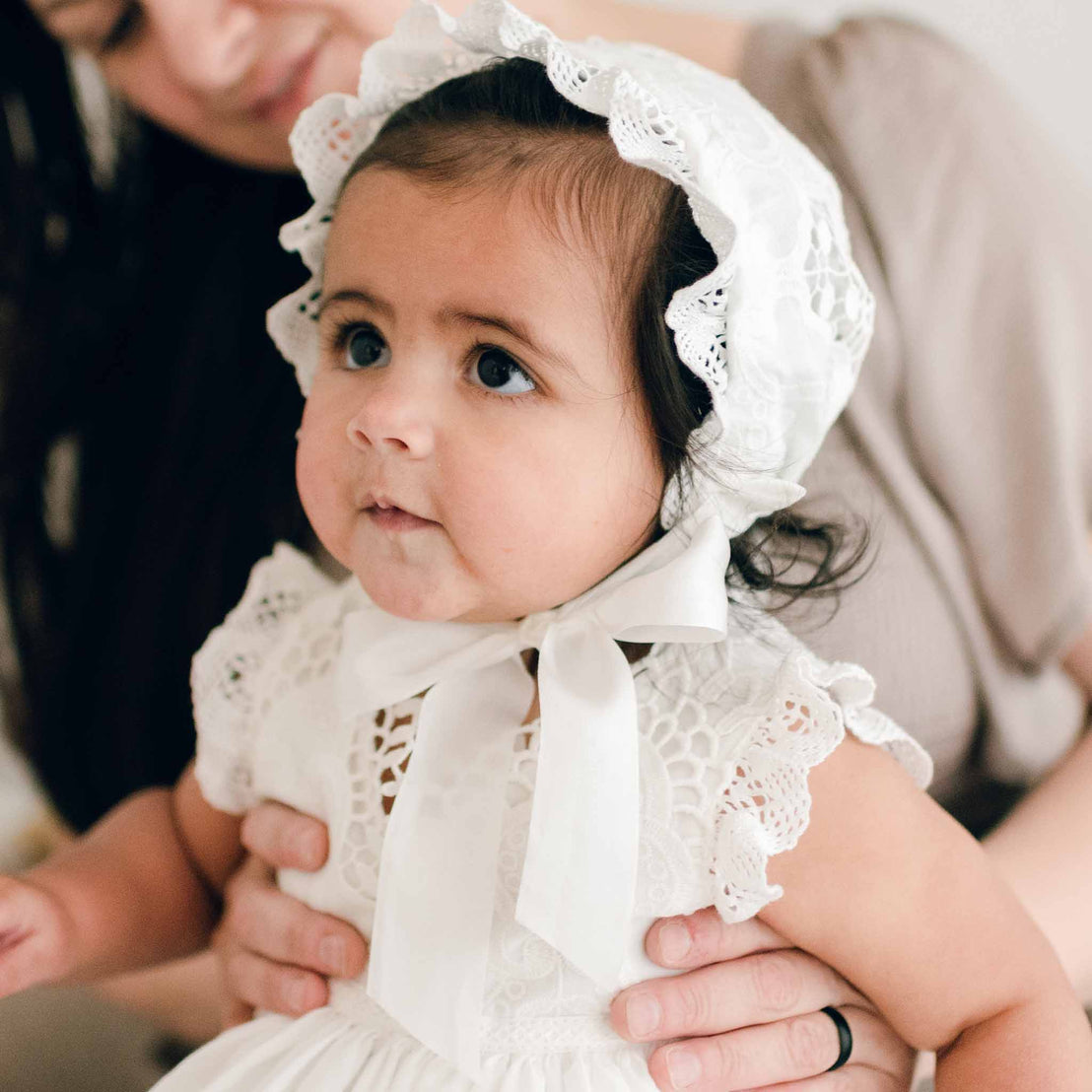 Baby girl with dark hair wearing a Lily Bonnet and matching lace dress looks to the side as her mother holds her.
