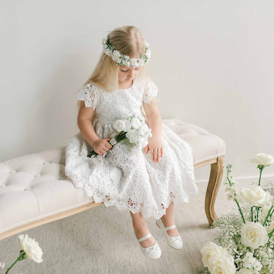 A young girl wearing the Lola Girls Lace Dress and a white floral crown, seated on a bench while looking down at a bouquet of white flowers.
