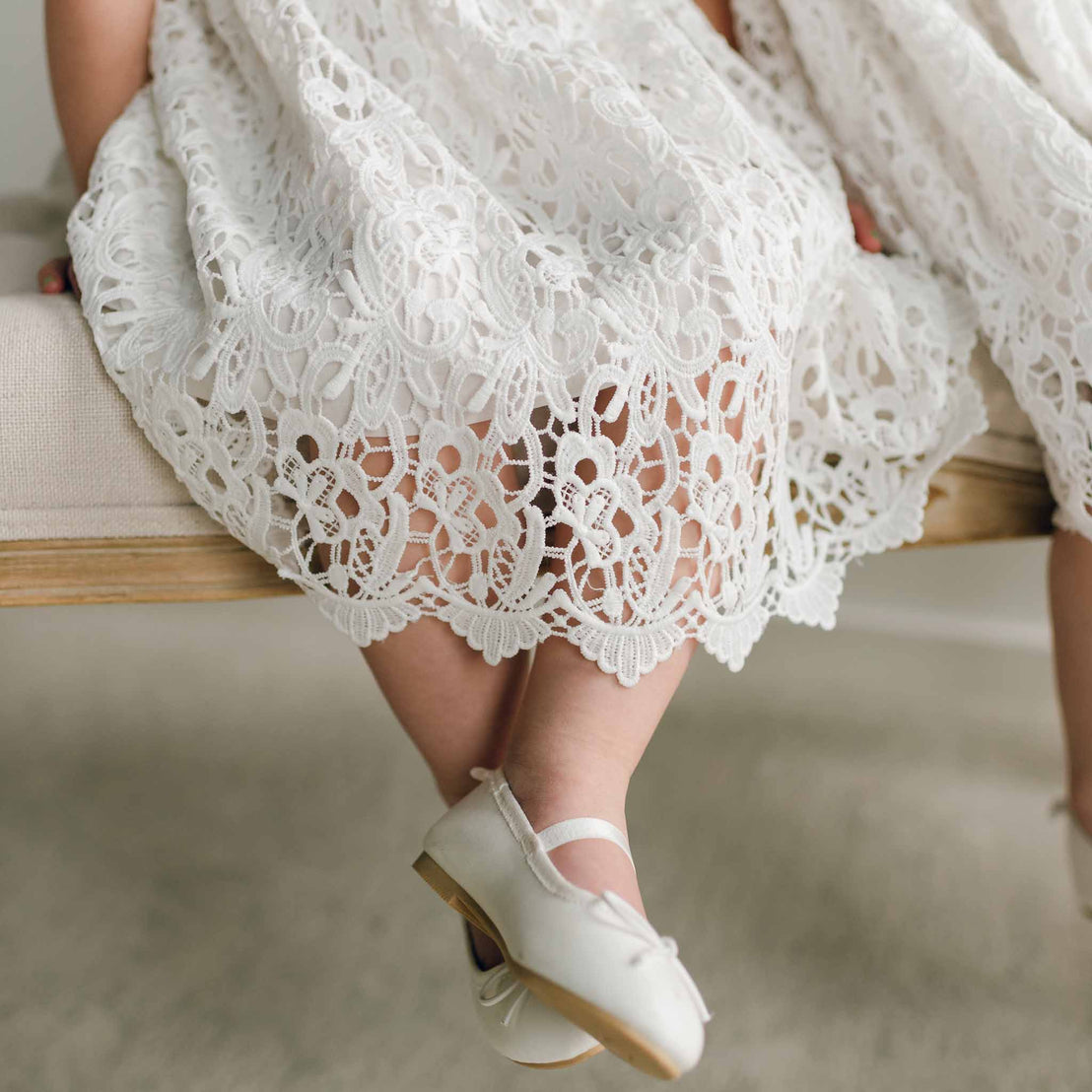 Detailed view of the lace hem of the Lola Girls Lace Dress and white shoes worn by a seated girl.