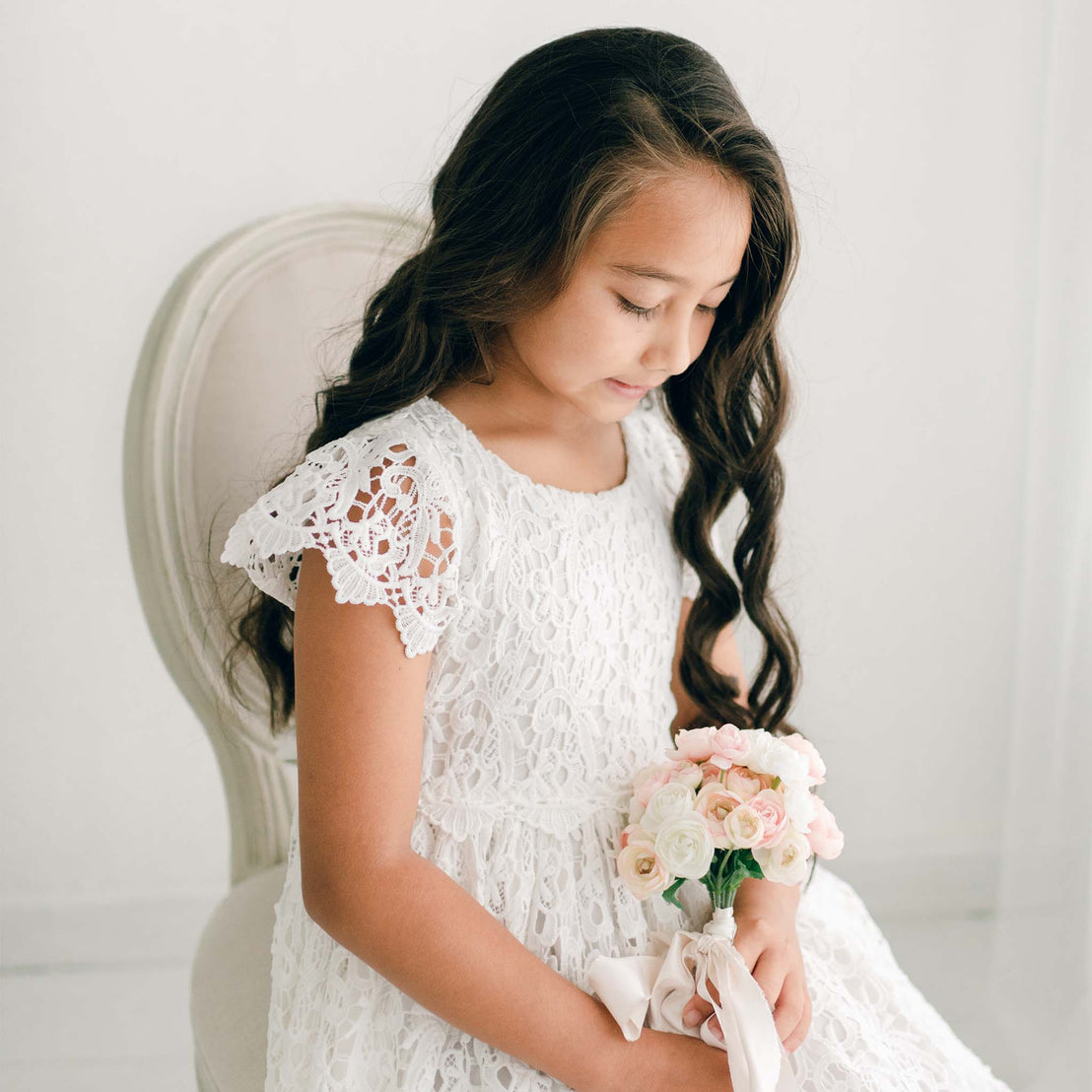 Side view of a girl wearing the Lola Girls Lace Dress with a lace headband, holding a small bouquet of blush and white flowers.