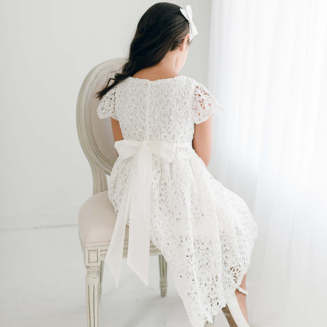 Back view of the Lola Girls Lace Dress on a girl seated in a chair, highlighting the white sash tied into a bow.