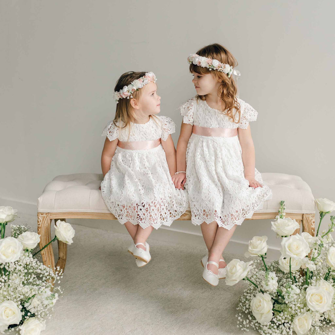 Full view of two young girls in the Lola Girls Lace Dress with pink satin sashes, seated side by side on a bench with floral arrangements nearby.