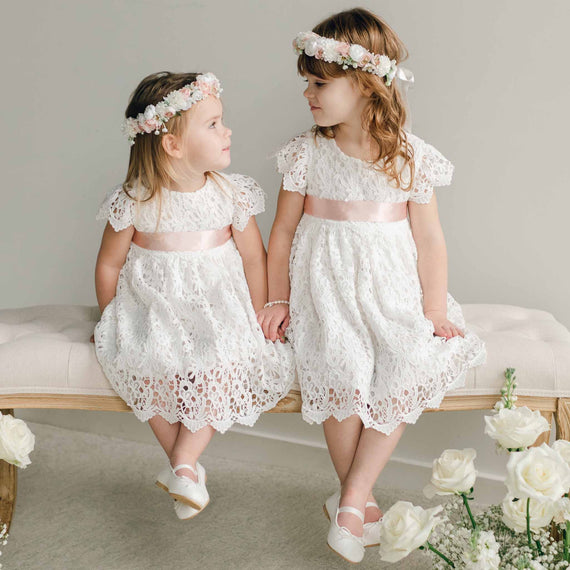 Two young girls wearing the Lola Girls Lace Dress with pink satin sashes and floral crowns, sitting on a bench surrounded by white roses.