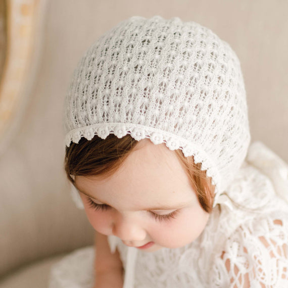 Close-up of a baby girl gazing down and wearing the Lola Knit Bonnet, made from soft light ivory knit acrylic with delicate cotton lace trim and silk ribbon ties.