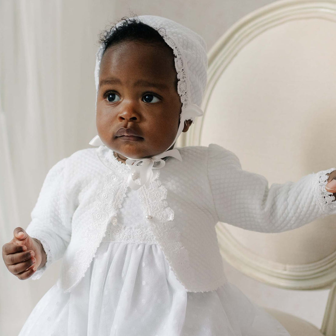 Baby in a white cotton sweater with a bonnet sitting on a chair.