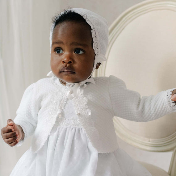 Baby in a white cotton sweater with a bonnet sitting on a chair.