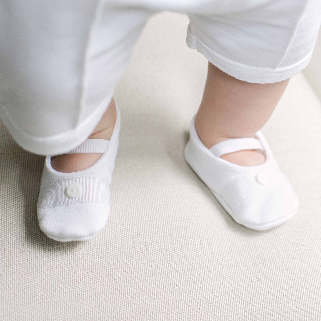 White knit baby booties with small white buttons, shown on a baby's feet wearing white pants.