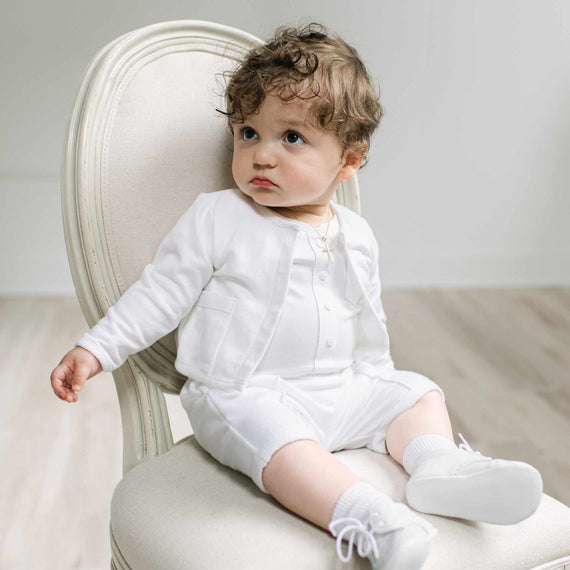 A baby with curly brown hair sits on a cream-colored upholstered chair, wearing a white long-sleeve cardigan, matching white shorts, white socks, and white shoes.