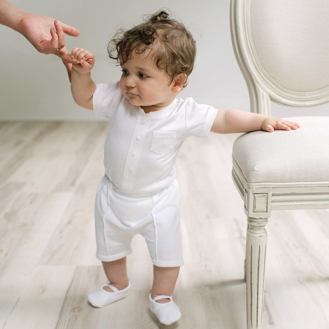 A baby wearing a white short-sleeve button-front romper and matching white shorts, standing on a light wood floor and holding an adult's hand next to a white upholstered chair.