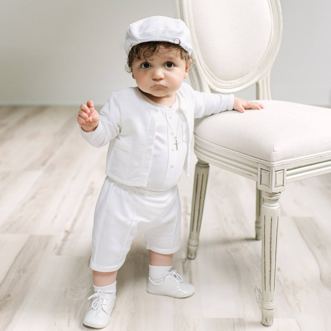 A baby standing on a light wood floor next to a white upholstered chair, wearing a white outfit with a matching white cap and white shoes.