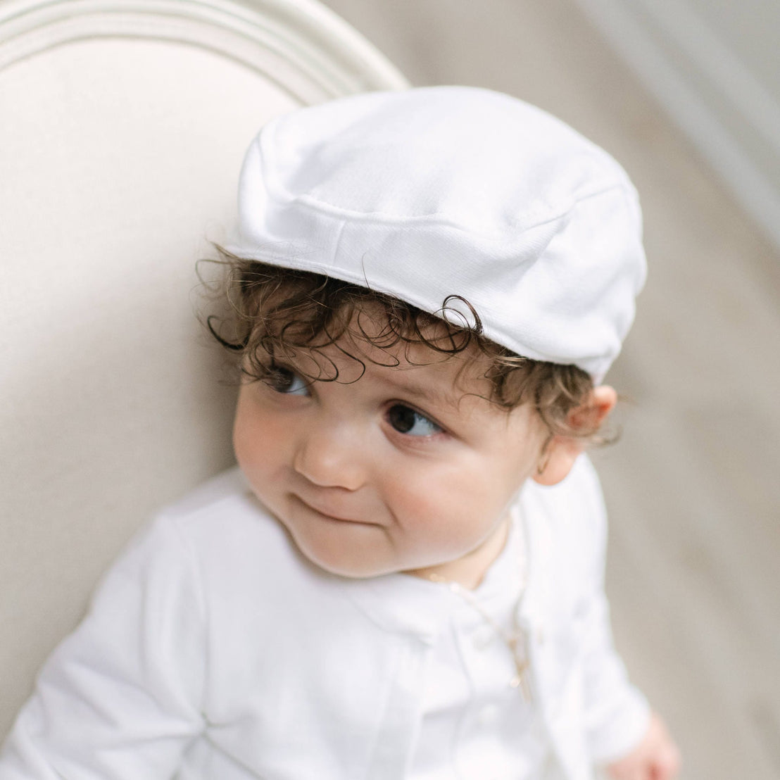 A baby wearing a white fabric cap and white outfit, sitting against a cream-colored upholstered chair.