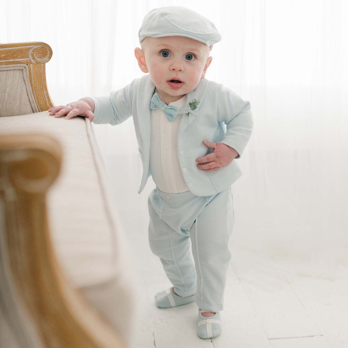 Baby boy standing in robin's egg blue suit, bow tie, newsboy cap and booties. Holding chair.