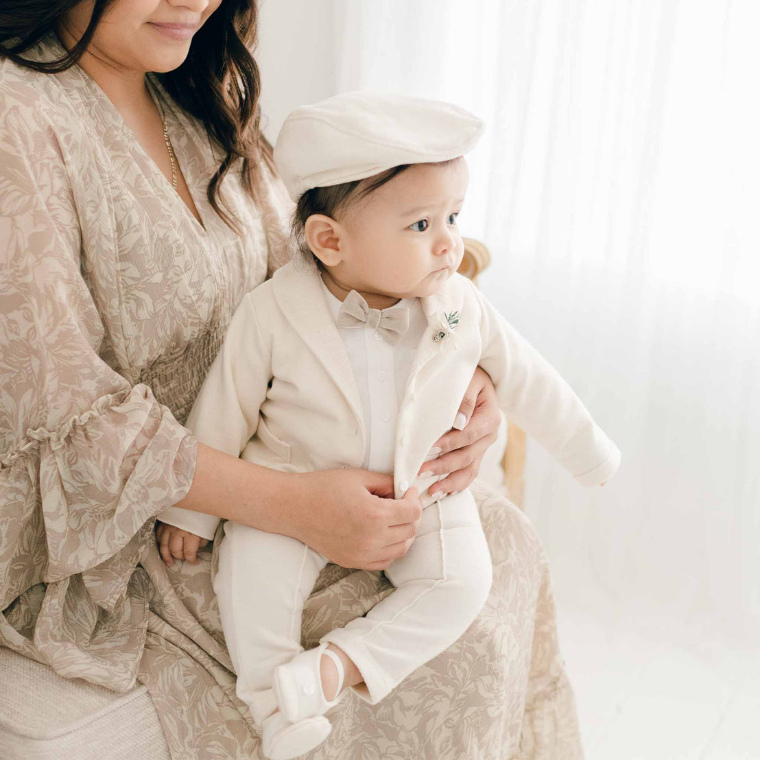 A baby wearing the Milo Tan 3-Piece Suit sits on a woman's lap, looking to the side, dressed in a jacket, bow tie, and cap.