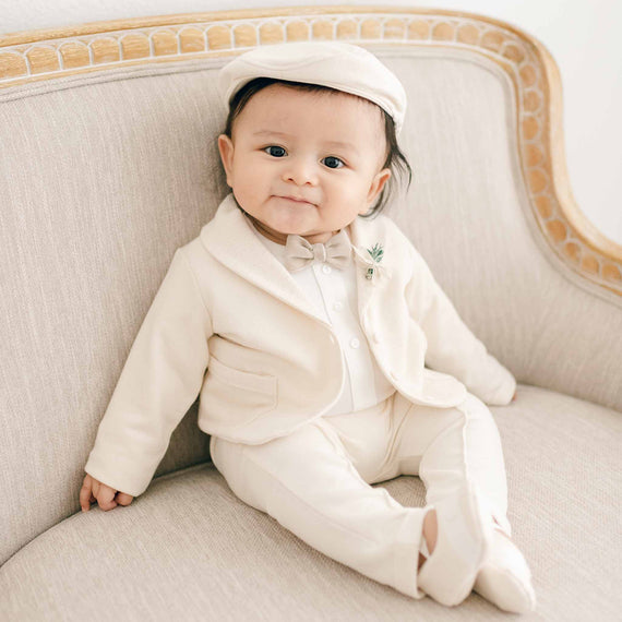 A baby wearing the Milo Tan 3-Piece Suit sits on a vintage-style couch, dressed in a jacket, bow tie, white shirt, and matching tan pants with matching booties.
