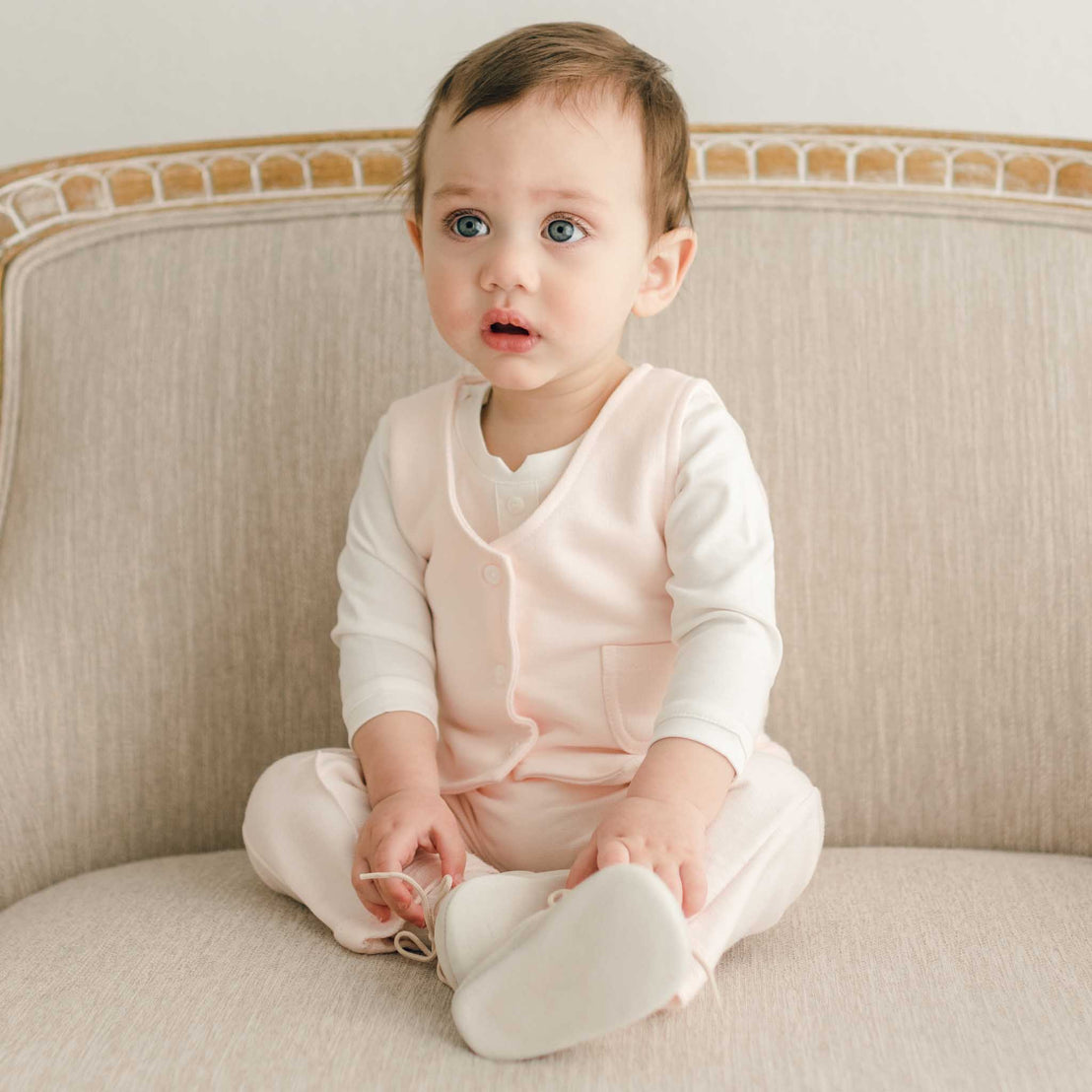 A baby sitting on a couch wearing the Milo Blush Vest over a long-sleeve shirt, paired with matching pants and shoes.
