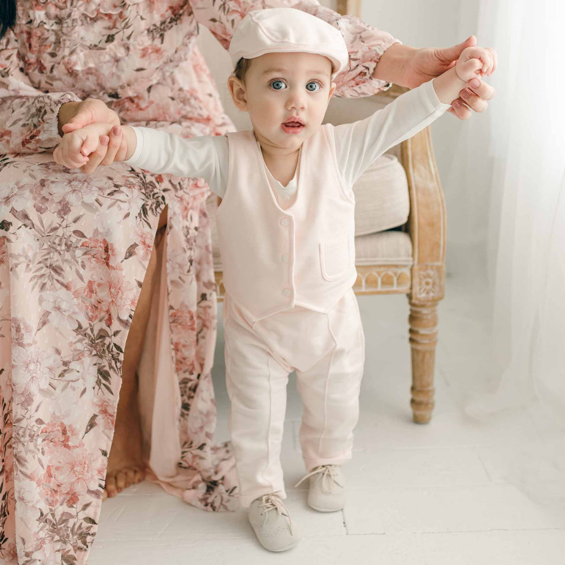 A baby wearing the Milo Blush Vest, standing with support while holding hands, paired with matching pants and a cap.