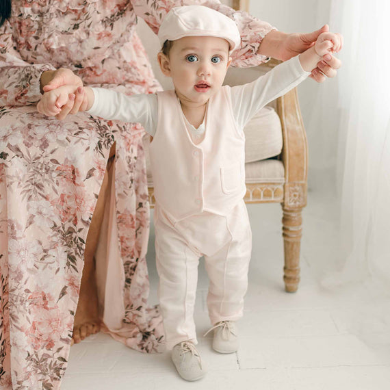 A baby wearing the Milo Blush Vest, standing with support while holding hands, paired with matching pants and a cap.