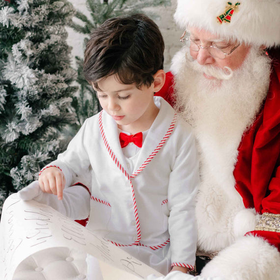 Child in a white outfit with red trim and bow tie sitting next to Santa Claus against a Christmas tree background.