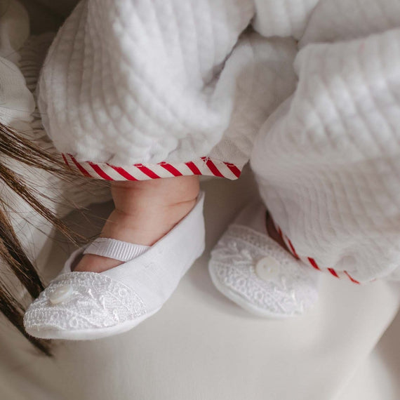 A baby wearing white clothing with red-striped cuffs is cradled in someone's arm. The baby's feet are adorned with delicate Nicky Booties, featuring a small button and Venice lace detailing.