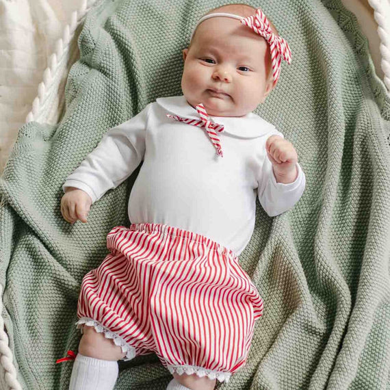 A baby lying in a basket, dressed in the Nicole Bloomer Set featuring red and white striped bloomers with a matching bow headband, ready for holiday events.