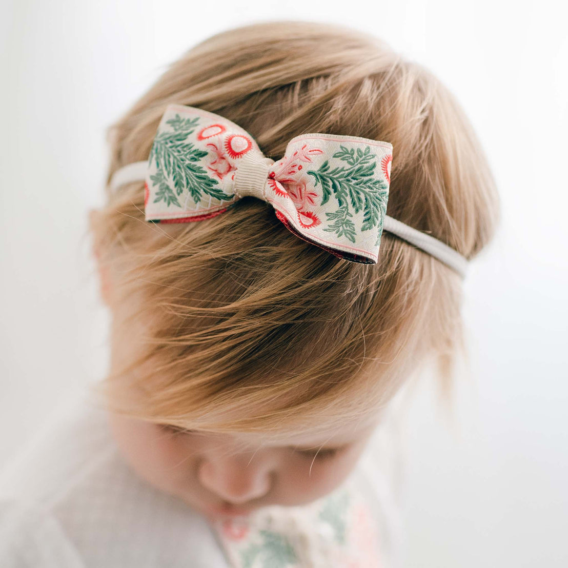 A young child with blonde hair is seen wearing a Noelle Headband, beautifully adorned with a decorative floral bow showcasing green foliage and pink flowers. Handmade in the USA, this enchanting accessory harmonizes perfectly with the gentle lighting as the child gazes downward.