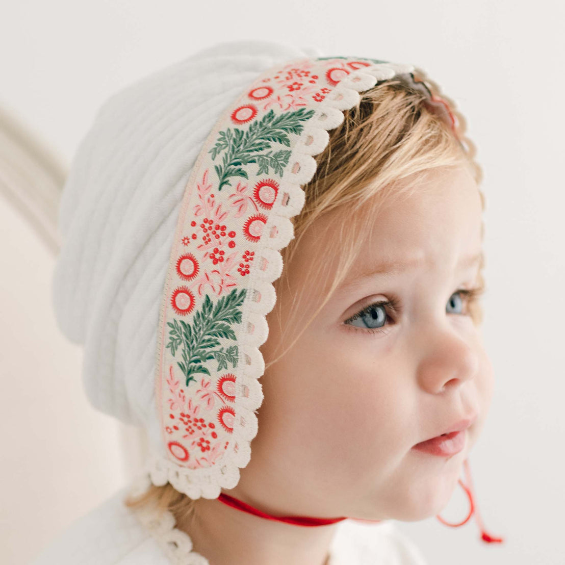 A young child wearing the Noelle Quilt Bonnet, featuring pink and green floral patterns and a delicate lace trim, complements their bright blue eyes and blonde hair as they gaze to the side. The 100% cotton bonnet sits comfortably as a soft, out-of-focus light background evokes a cozy holiday season feeling.