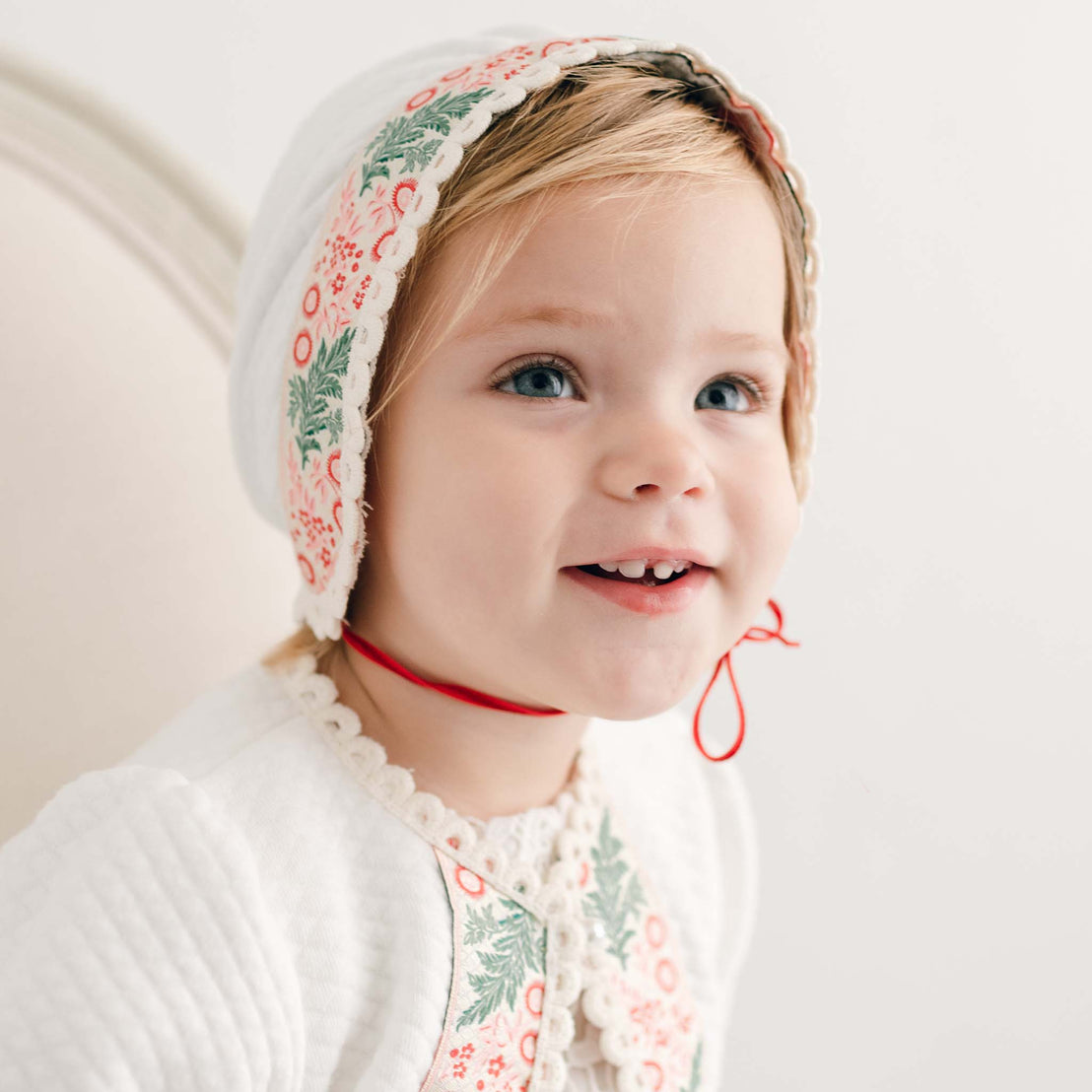 A smiling toddler wears the Noelle Quilt Bonnet along with a white outfit adorned with floral embroidery, radiating festive cheer. The child is seated, facing slightly to the side against a light-colored background, ideal for capturing the holiday season spirit.