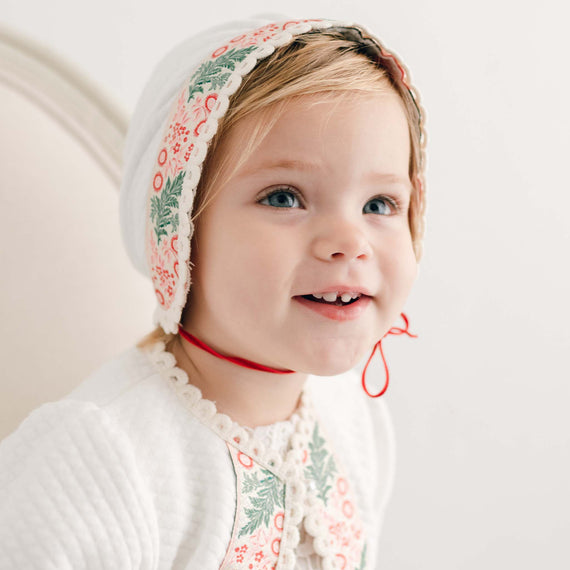 A smiling toddler wears the Noelle Quilt Bonnet along with a white outfit adorned with floral embroidery, radiating festive cheer. The child is seated, facing slightly to the side against a light-colored background, ideal for capturing the holiday season spirit.