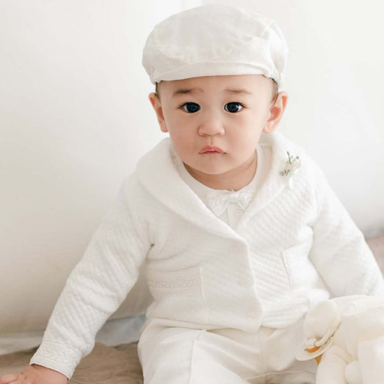 Baby sitting on a bed wearing the Oliver Quilted Jacket Suit with a matching cap and white stuffed bunny resting on their lap.