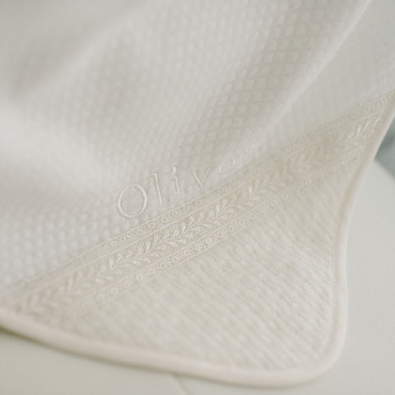 A close-up image of the ivory Oliver Personalized Blanket featuring the ivory, textured cotton fabric with delicate embroidered detailing. The embroidery includes the name "Oliver" and intricate, leafy patterns along the trim.