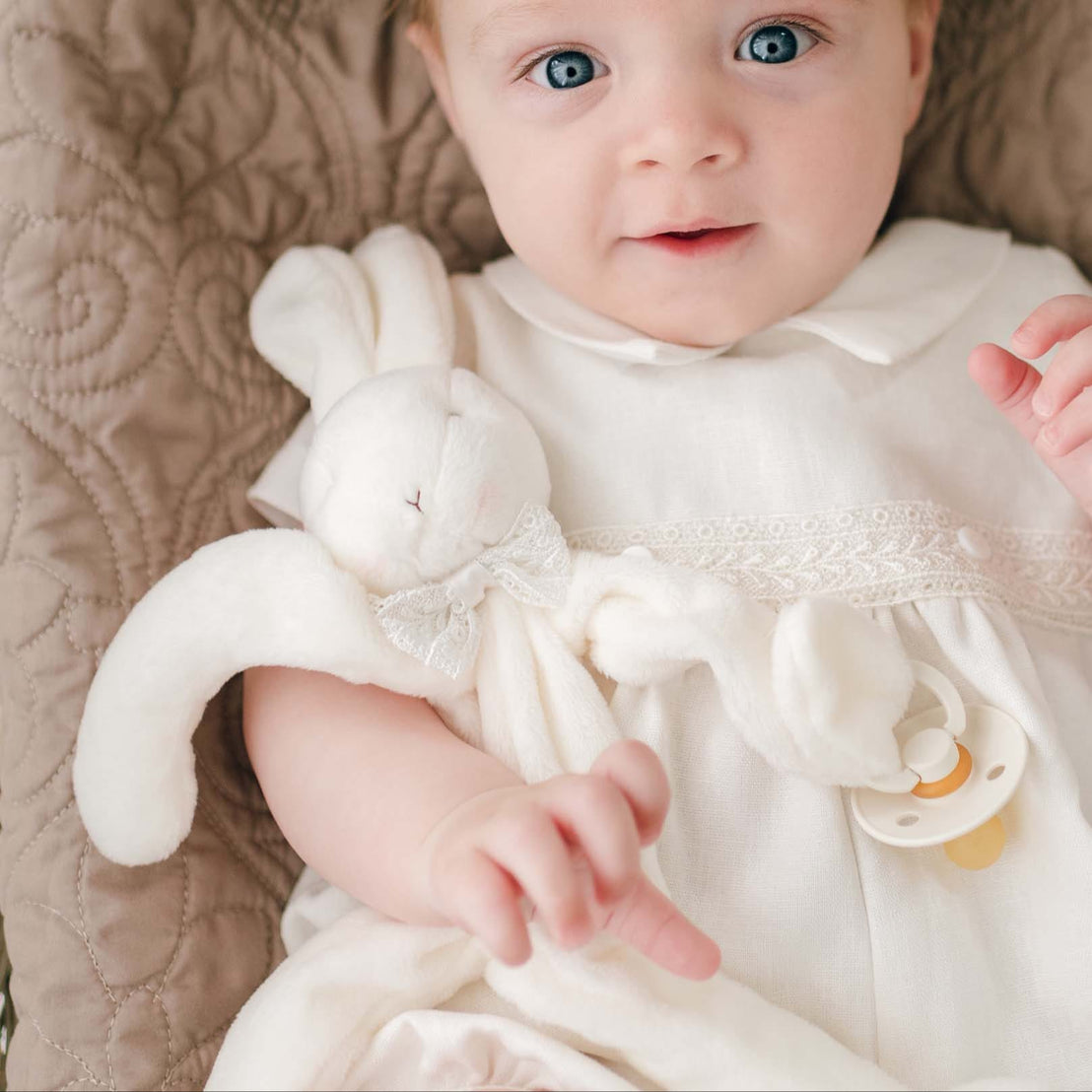 A baby with light brown hair and blue eyes lies on a quilted beige blanket, wearing the Oliver Romper in white. The baby holds an Oliver Silly Bunny Buddy | Pacifier Holder made of soft white velour and has a baby-safe pacifier clip attached to its hand. The baby has a calm, curious expression.