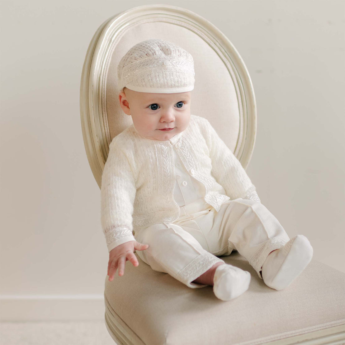 A baby sitting on a vintage-style chair dressed in the Oliver Sweater Pants Suit, showcasing the detailed knit sweater, lace-trimmed pants, and matching cap.