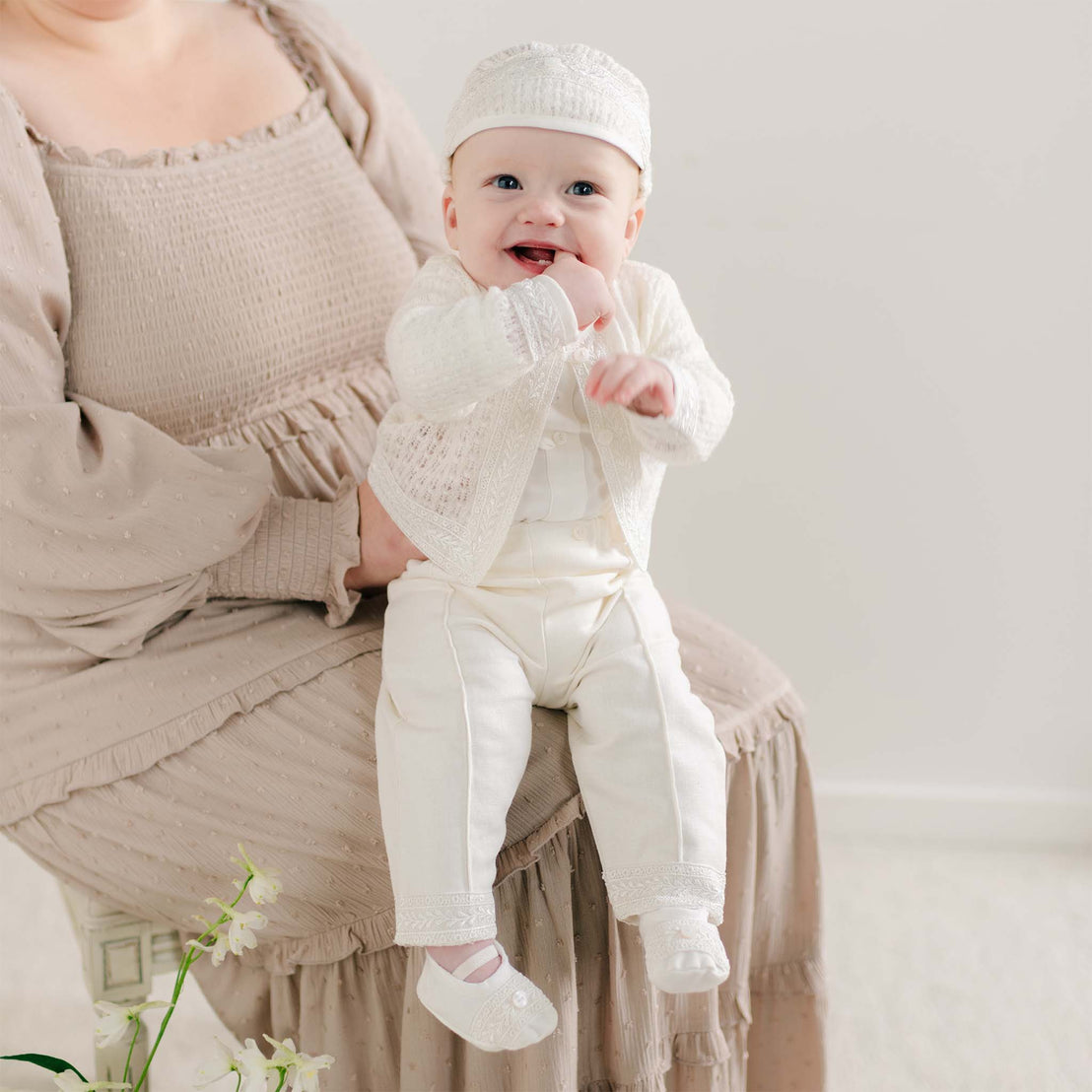 A baby sitting on a parent's lap wearing the Oliver Sweater Pants Suit in soft ivory, featuring a knit sweater with delicate lace trim, matching pants, and a coordinating cap.