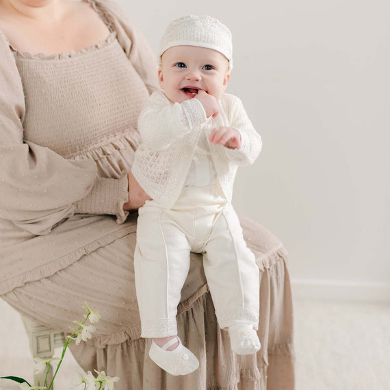 A baby sitting on a parent's lap wearing the Oliver Sweater Pants Suit in soft ivory, featuring a knit sweater with delicate lace trim, matching pants, and a coordinating cap.